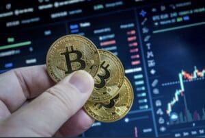 Ruka drži zlatne Bitcoin novčiće uz zamućen ekran s podacima o trgovanju.