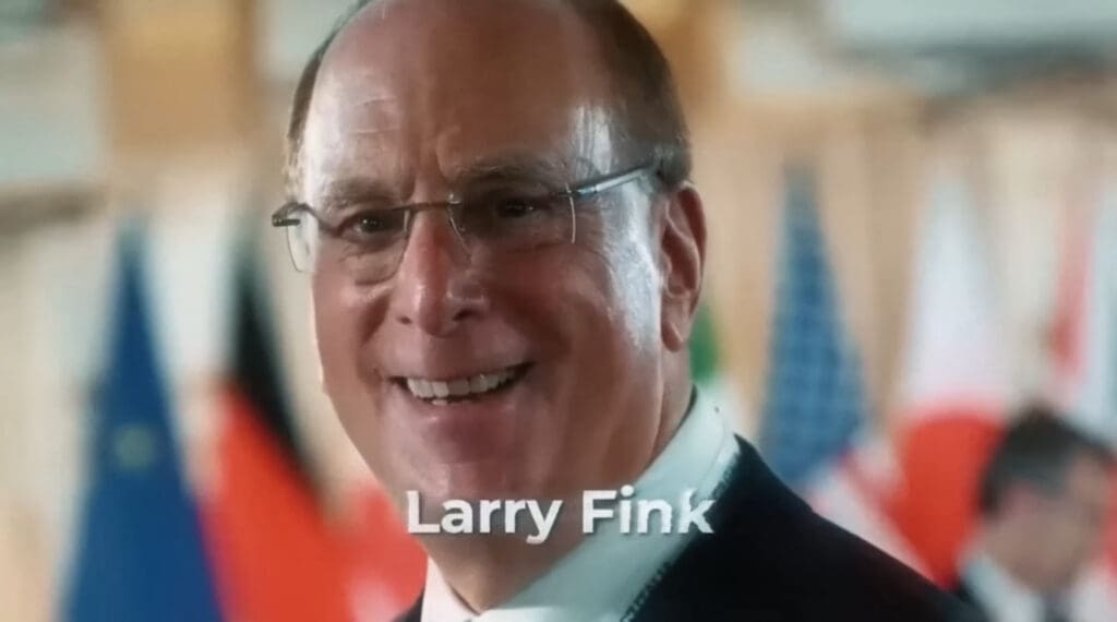 Larry Fink na međunarodnom događaju ispred zastava, nosi odijelo i naočale.